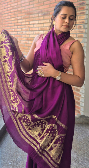 Woman in a purple saree with gold patterns standing indoors.